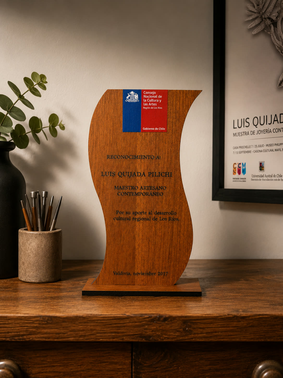 Galardón de madera con forma ondulada sobre un mueble, grabado con el reconocimiento del Consejo Nacional de la Cultura y las Artes a Luis Quijada Pilichi.