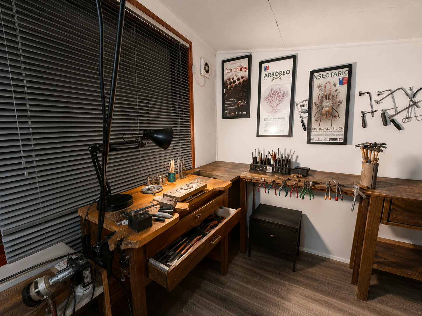 Taller de joyería en Valdivia: banco de trabajo de madera con herramientas de mano, alicates colgados en una barra y los afiches de Reino Fungi, Arbóreo e Insectario enmarcados en la pared.